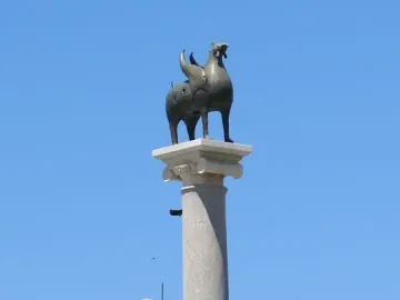 Pisa Griffin statue on a tall stone column above the roof of a cathedral, against a blue sky.