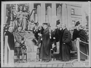 Alma Mater, a bronze seated woman with a laurel wreath, scepter, and open book, on a marble pedestal surrounded by people.