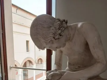 Boy with Thorn sculpture seen in profile, a light-colored boy leaning forward with his hands around his raised foot near a window.