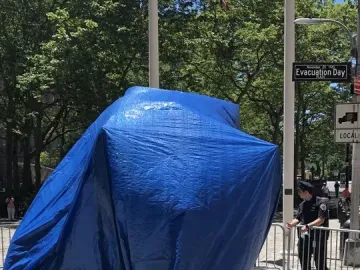 Charging Bull covered by a blue tarp on cobblestone ground, with police barriers and a street sign in the background.