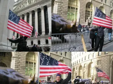 Charging Bull sculpture on a city street behind metal barricades, with people nearby holding American flags and classical buildings in the background.