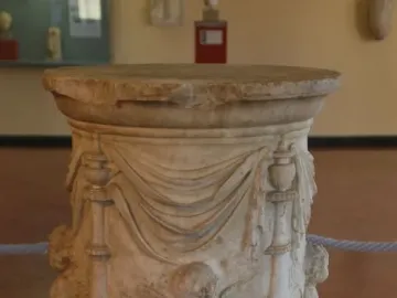 Round marble Four Seasons Altar with carved drapery, decorative pillars, and a winged putto holding grapes and a basket of fruit.