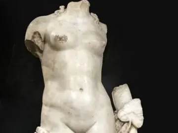 Headless white stone sculpture of Venus Unica on a metal base, with broken limbs and draped fabric against a dark background.