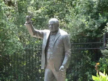 Winston Churchill bronze statue standing on a stone base and raising his right hand in a V for victory gesture while holding a cane.