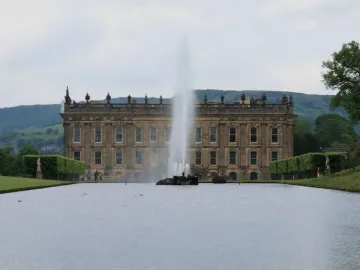 The Emperor Fountain shoots high in front of Chatsworth House, with green hills and trees under an overcast sky.