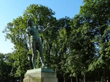 The Greek Actor sculpture on a tall stone pedestal in a park, with flowers and trees behind it.