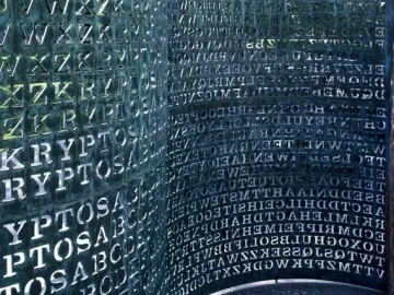 Kryptos, a curved metal sculpture covered with cut-out letters and numbers, with patterned shadows cast on the ground.