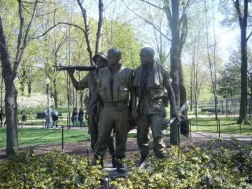 The Three Soldiers bronze sculpture standing in a park with trees, grass, and flowering bushes nearby.