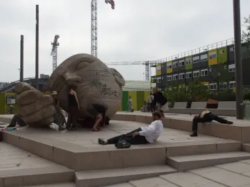 Écoute sculpture showing a large stone head lying on its side beside a cupped hand, with people around it in a plaza.