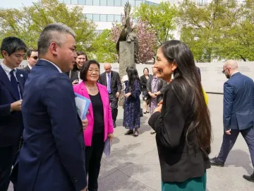 People gathered on a sunny plaza near the Japanese American Memorial to Patriotism During World War II, with a tall metallic sculpture and stone walls in the background.