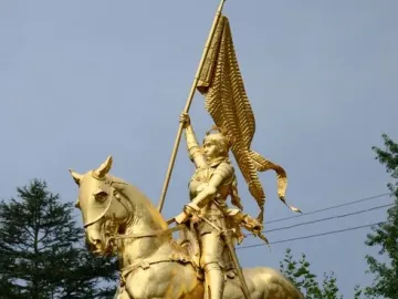 Golden equestrian statue of Joan of Arc in armor holding a flag-topped staff on a stone pedestal.