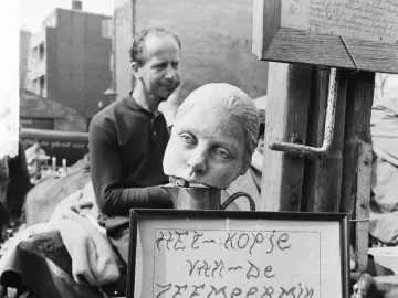 Detached head sculpture of The Little Mermaid resting in a cup, with a framed Dutch sign below it and a man seated in the background.