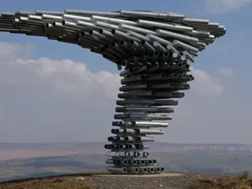 Singing Ringing Tree sculpture made of stacked silver metal pipes on a hillside with rolling landscape and distant wind turbines.