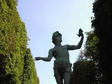 The Greek Actor statue on a stone pedestal with one arm raised holding a mask, set along a tree-lined path with a domed building in the distance.