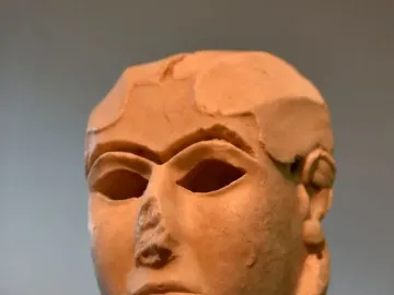 The Warka Mask, a light-toned ancient stone head with stylized facial features and hollow eye sockets on a dark pedestal.