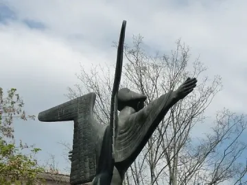 The Angel of Peace bronze statue with abstract wings and flowing robes, shown outdoors against bare trees and a cloudy sky.