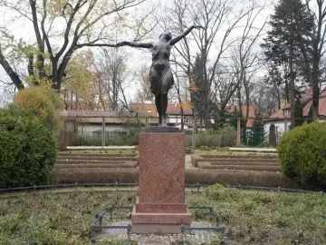 The Dancing Girl statue on a stone pedestal in a park with bushes and bare trees nearby.