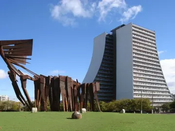 Monument to the Azoreans on a green lawn with a tall white building behind it under a blue sky.