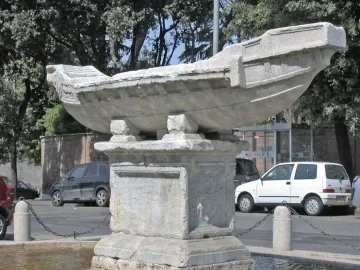 Fontana della Navicella stone boat fountain on a pedestal in a shallow basin, with trees and parked cars in the background.