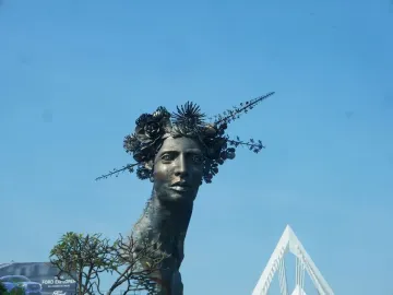 Reminiscencia sculpture showing a dark metal woman\'s head with an elaborate crown of flowers and branches against a blue sky.