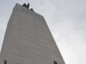 This is the Place Monument with bronze figures at its base and a tall stone shaft rising into the cloudy sky.
