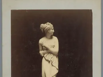 Sepia photograph of the Venus Italica marble statue, shown full-length against a dark background.