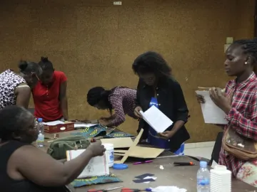 Women gathered around a table at a cartonería workshop, working with cardboard, fabric, scissors, and glue.