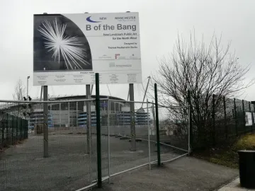 Information board for B of the Bang beside fenced gravel ground and a stadium building.