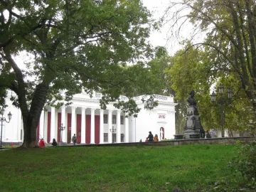 Pushkin bust on an ornate pedestal in a park with trees, a lamppost, and a columned building behind it.