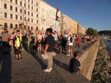 People gathered beside the Shoes on the Danube Bank memorial along a riverbank lined with shoe sculptures.