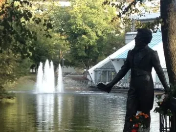 Pushkin statue on a stone pedestal near a pond with fountains, with trees and a modern building in the background.