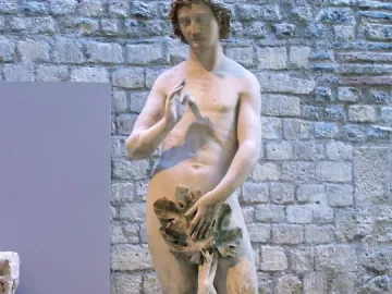 Adam statue holding leaves over his groin beside a stone wall and tree trunk.