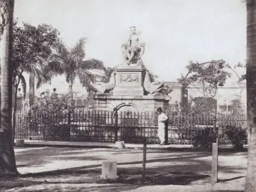 Fuente de la India stone fountain with a seated woman statue, carved sea creatures, and a surrounding iron fence.