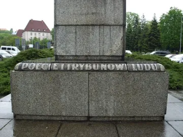 Lower section of the Mickiewicz Monument with a grey granite pedestal and carved inscription, surrounded by bushes and wet paving stones.