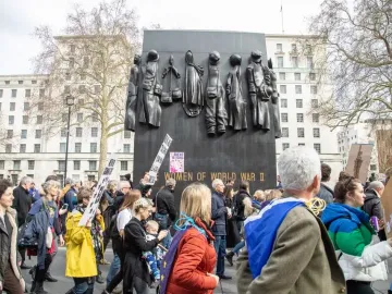 The Monument to the Women of World War II stands behind a crowd of protesters in a city square.