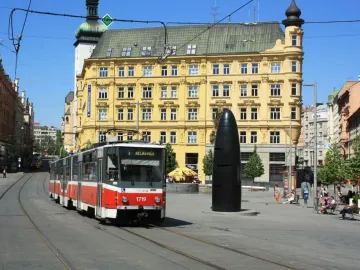 The Brno astronomical clock stands in a city square as a tram passes in front of it, with historic buildings in the background.
