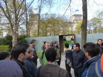 Translucent text panels and a dark metal gateway at the Memorial to the Murdered Sinti and Roma of Europe, with people gathered nearby.