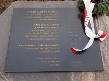 Dark grey plaque on the Monument of Gratitude to the Iranian People with gold Polish and Persian inscriptions and a white-and-red ribbon draped over one side.