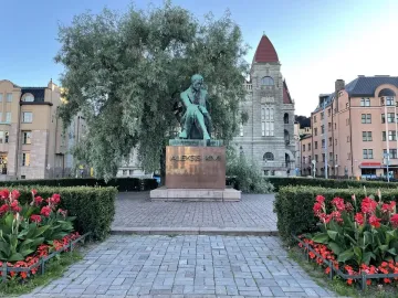 Aleksis Kivi Memorial bronze statue of a seated figure on a pedestal in a paved square, with hedges, red flowers, and buildings behind it.
