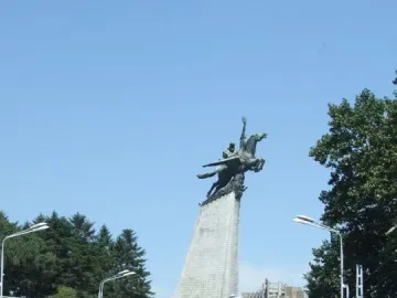 The Chollima Statue rises above a multi-lane road in Pyongyang, with trees, streetlights, and buildings in the background.