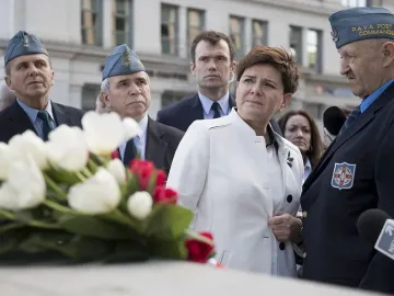 People gathered at the Katyń Memorial with white and red flowers in the foreground and a man in a blue uniform speaking into a microphone.