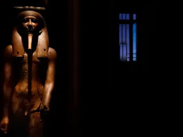 Wooden statue of King Hor with a pharaonic headdress and false beard on a black background.