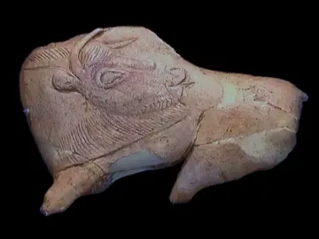 Carved bison sculpture with its tongue extended toward its flank, showing engraved fur details and a cracked, incomplete form.