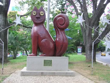 El Gato del Río cat sculpture on a stone pedestal in a park, with a small bird perched on its curled tail.