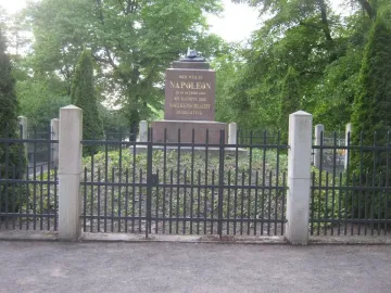 Napoleonstein stone monument behind a black metal fence in Leipzig, framed by green trees.