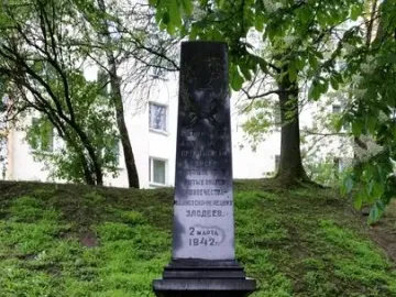 The Pit monument, a tall grey stone memorial with inscriptions and a carved emblem at the top, stands on a grassy hill with wreaths and flower baskets at its base.