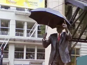 Allow Me bronze sculpture of a suited man holding an open umbrella and raising one hand on a brick plaza.