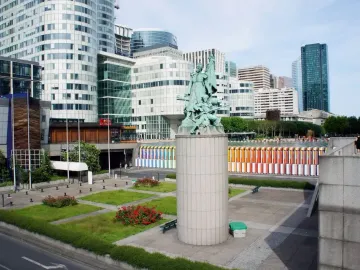La Défense de Paris sculpture on a tall pedestal in a modern urban plaza with lawns and skyscrapers.