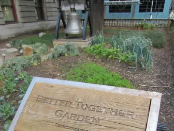 A large silver Liberty Bell display stands in a garden beside a Better Together Garden sign.