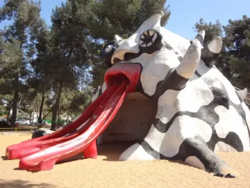 The Golem sculpture with a red mouth and three red slide chutes on sandy ground under a blue sky.